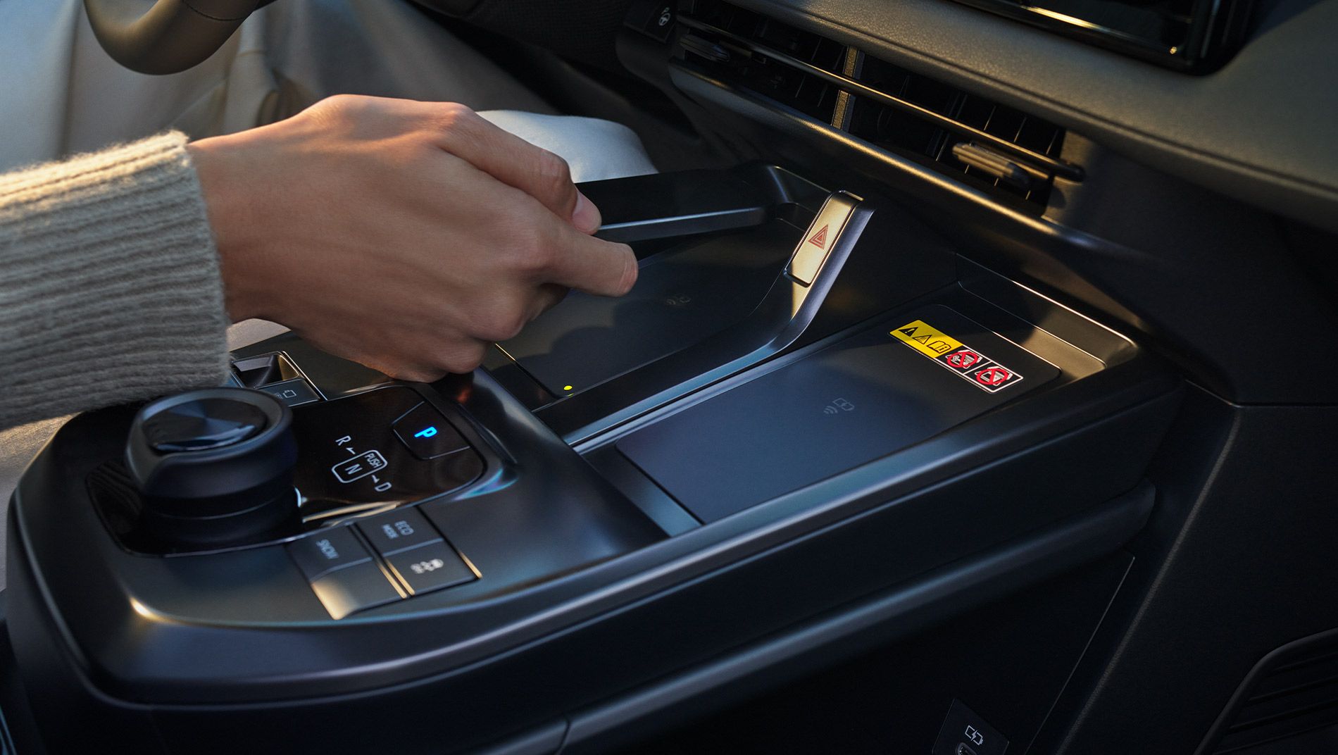 A person’s hand is gripping the gear selector in the center console of a modern car interior, surrounded by control buttons and a digital interface.