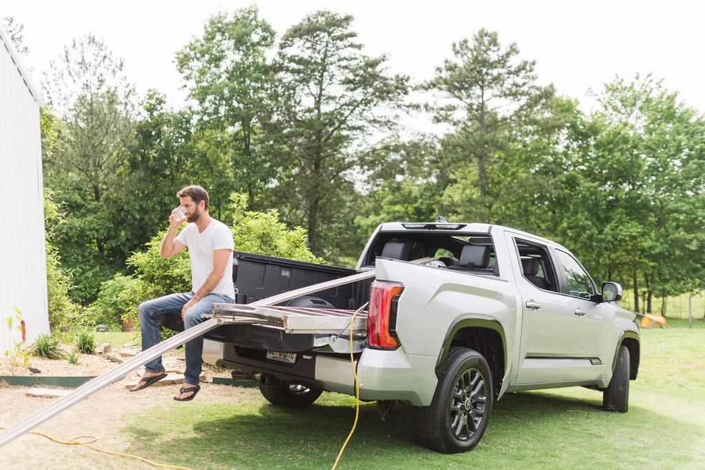 A man sits on the open tailgate of a silver pickup truck, drinking from a cup. He is wearing a white t-shirt, jeans, and sandals. The truck is parked on grass near trees and a white building.