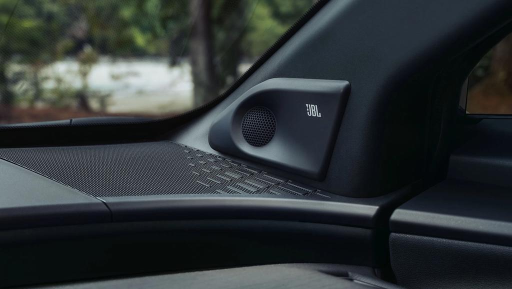 Close-up of a JBL speaker integrated into a car’s dashboard, with patterned detailing on the surface and a blurred view of trees outside the window.