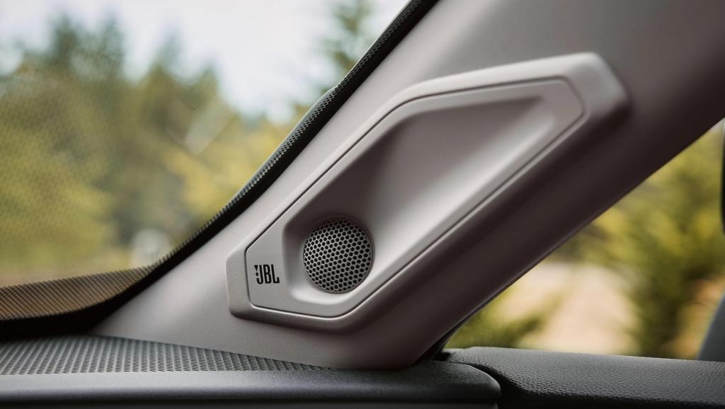 Close-up of a JBL car speaker mounted on the interior A-pillar near the windshield, with trees and greenery visible outside through the car window.