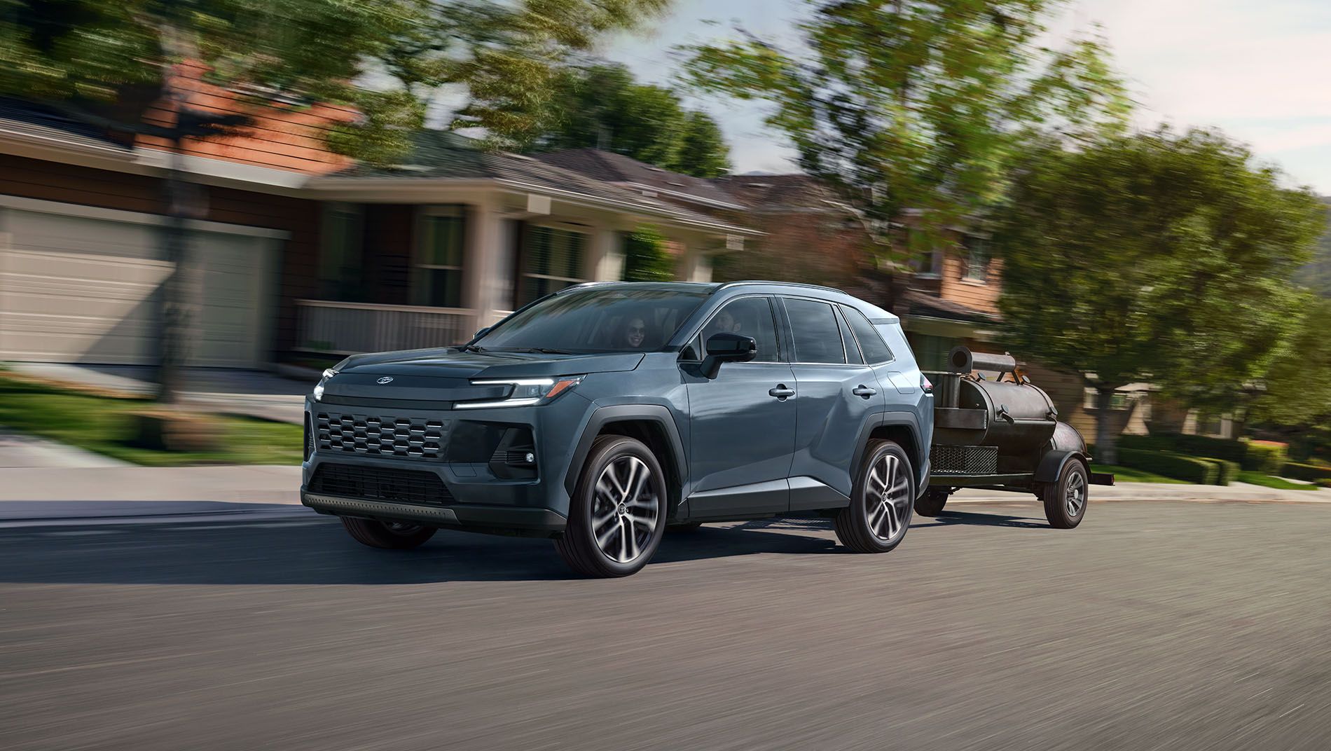 A gray SUV drives down a suburban street lined with houses and trees, towing a small black trailer behind it on a sunny day.