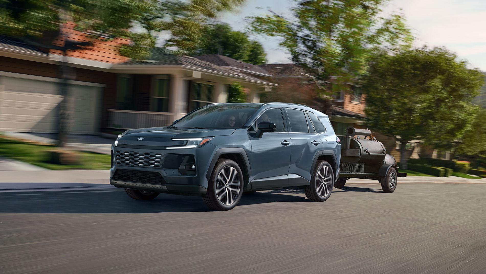 A gray SUV drives down a suburban street lined with houses and trees, towing a small black trailer behind it on a sunny day.