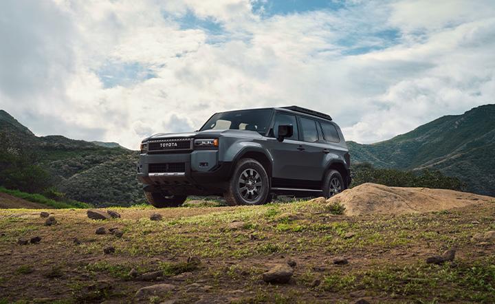 2024 Toyota Land Cruiser Hybrid parked on a mossy mountain ledge with rolling hills behind