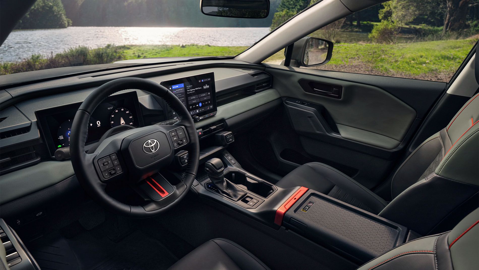 Interior of a modern Toyota vehicle featuring a black steering wheel, digital dashboard, touchscreen display, gear shifter, and leather seats with red accents. Outside, there is greenery and a lake visible through the windows.
