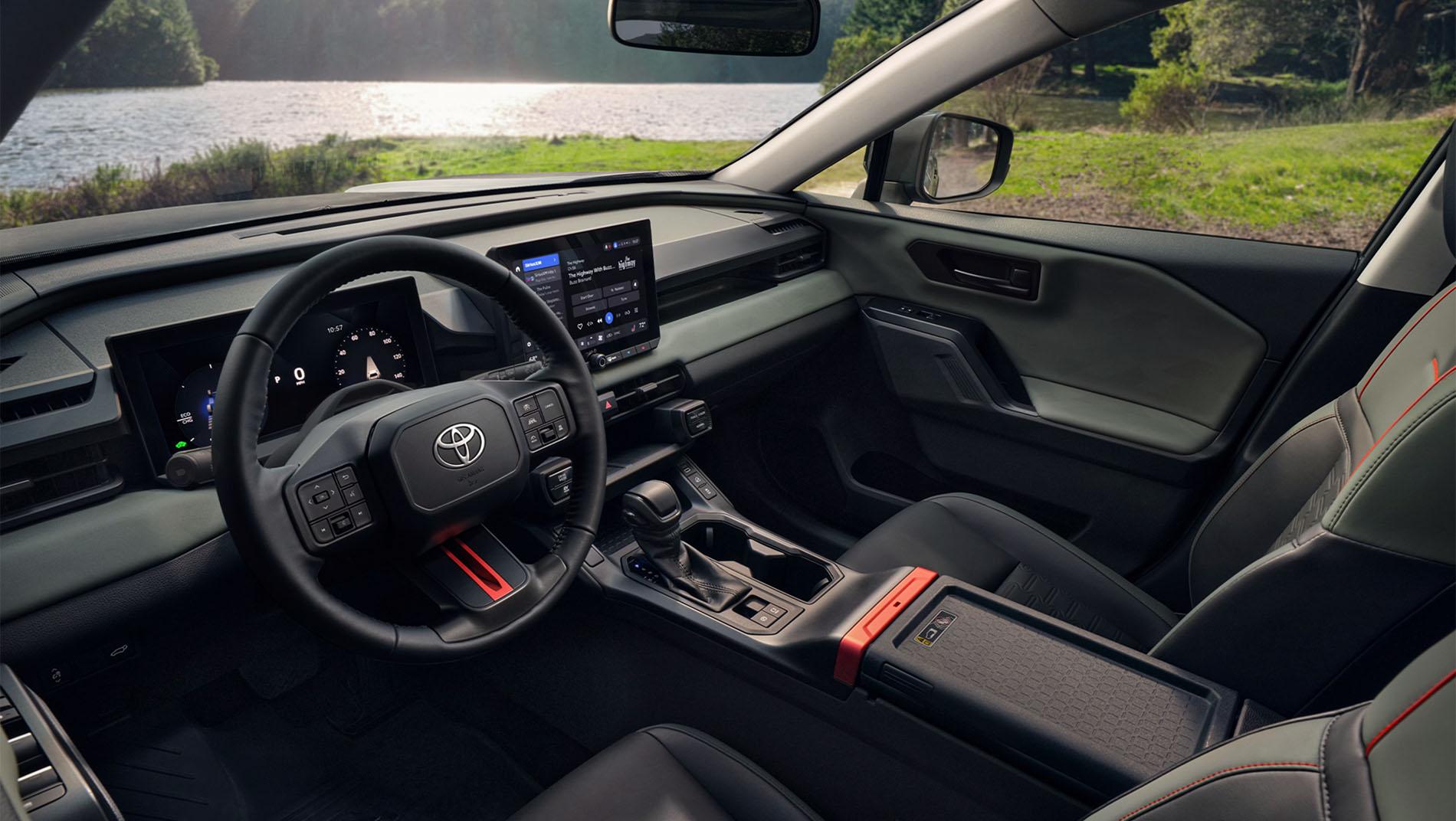Interior of a modern Toyota vehicle featuring a black steering wheel, digital dashboard, touchscreen display, gear shifter, and leather seats with red accents. Outside, there is greenery and a lake visible through the windows.