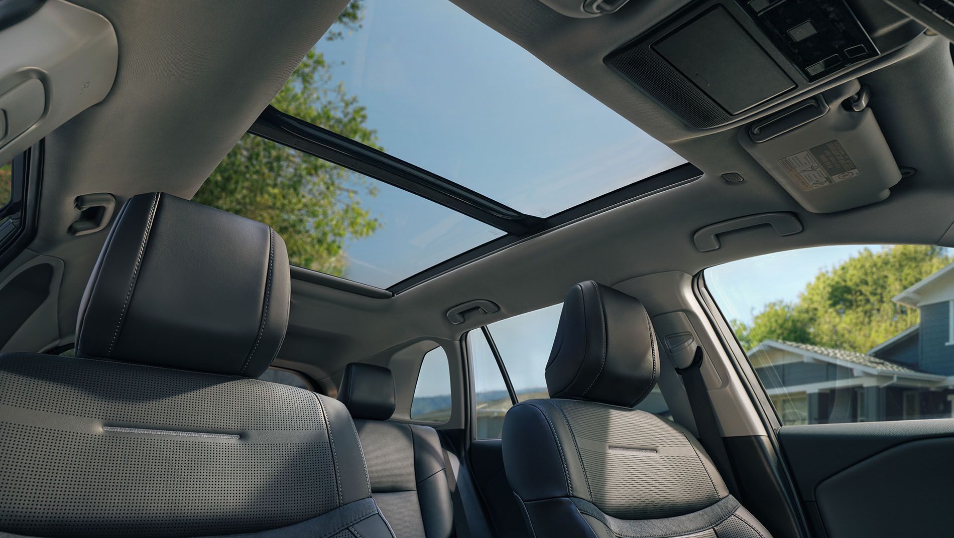 Interior view of a car showing two black front seats and an open panoramic sunroof. Blue sky, trees, and part of a house are visible through the roof, letting in natural light.