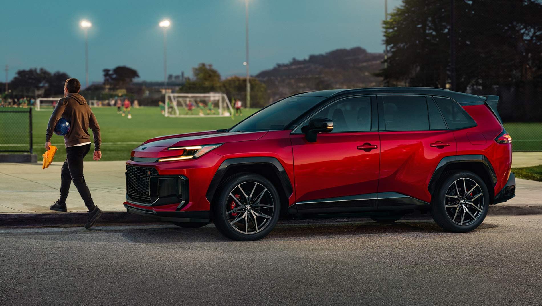 A red SUV is parked on the street beside a sports field where people are playing soccer. A person holding equipment walks on the sidewalk near the car at dusk.