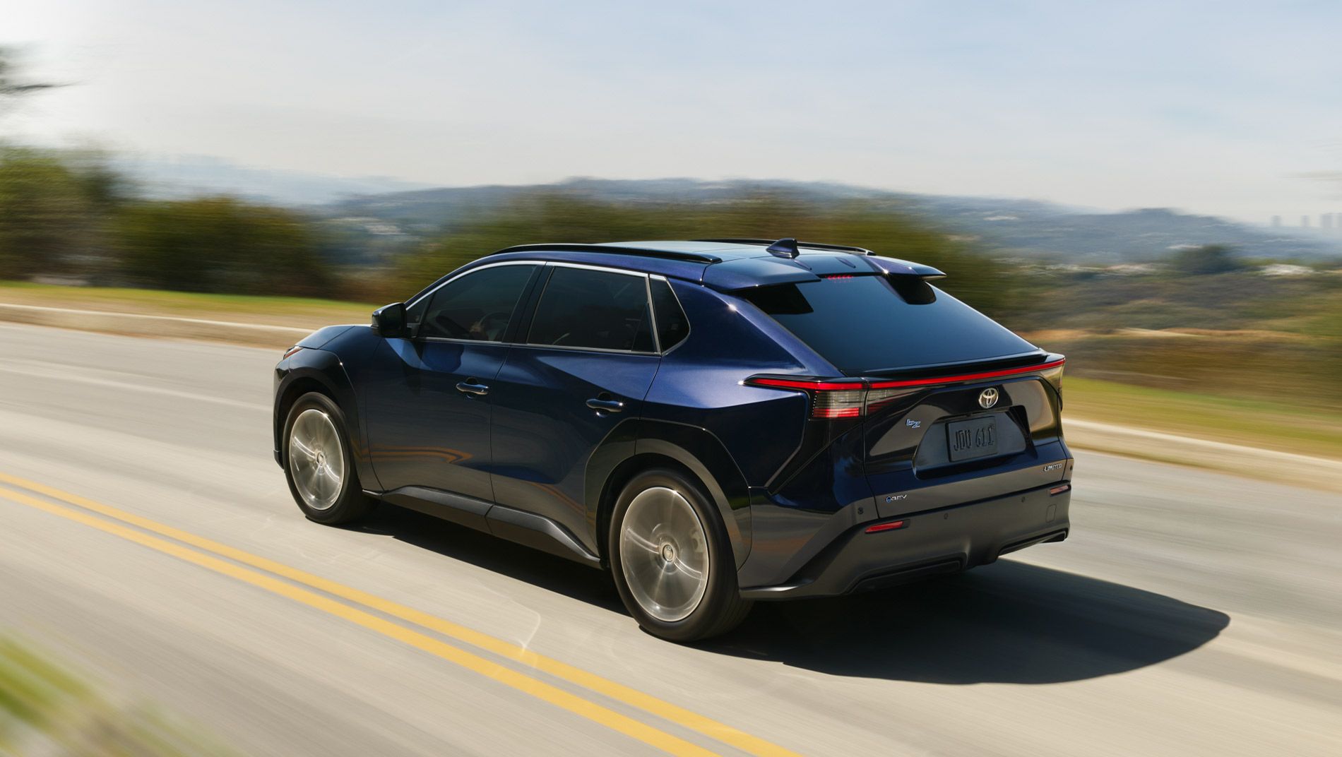 A dark blue Toyota SUV drives on a sunny, winding road with blurred trees and hills in the background, indicating motion. The vehicle is viewed from the rear left side.
