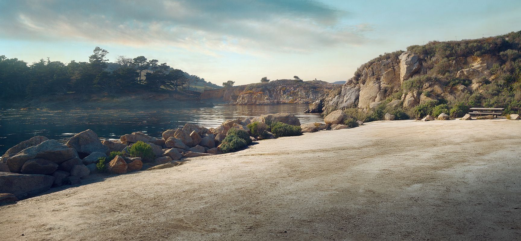 A calm lakeside scene with rocky shores, scattered bushes, and a picnic table. The water reflects the sky, and trees grow on the rocky hills in the background under a partly cloudy sky.
