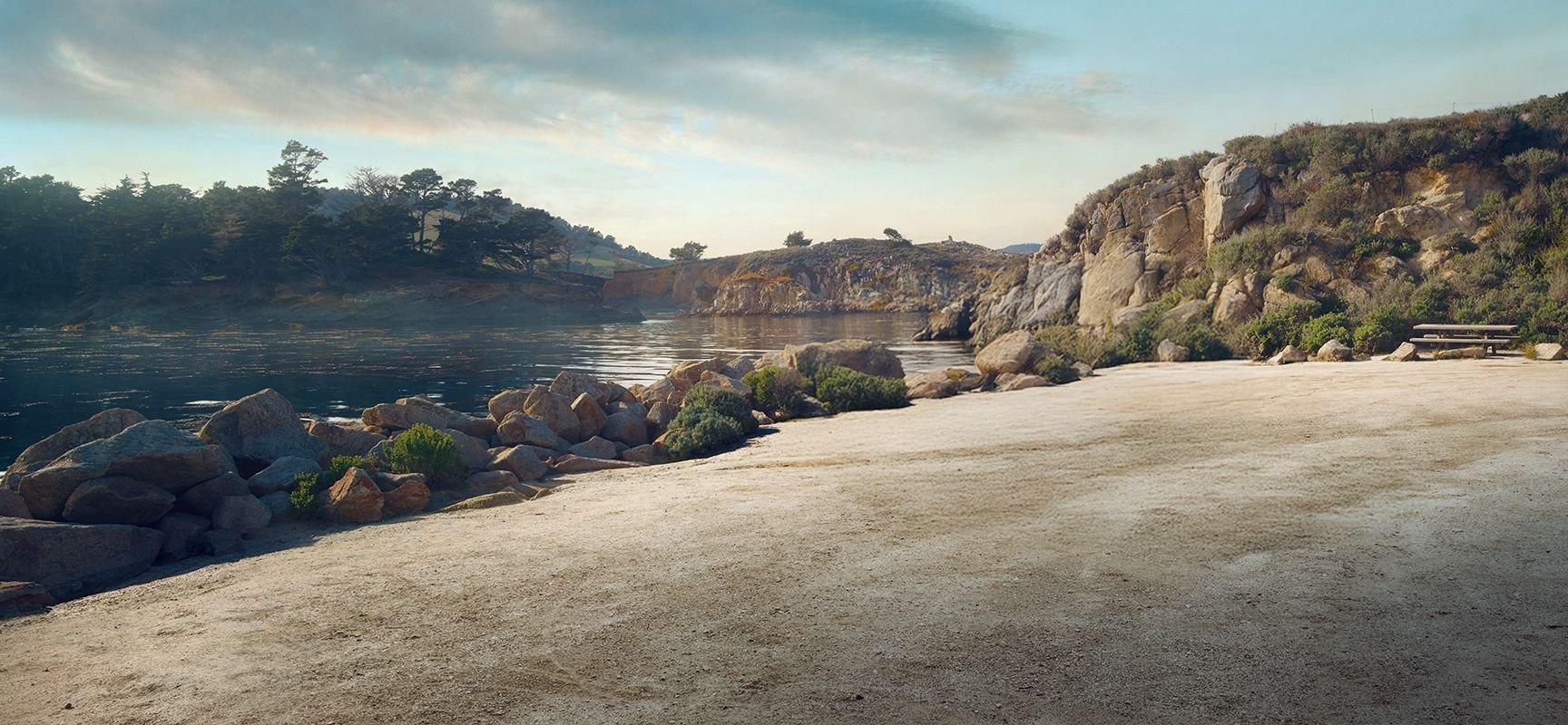 A calm lakeside scene with rocky shores, scattered bushes, and a picnic table. The water reflects the sky, and trees grow on the rocky hills in the background under a partly cloudy sky.