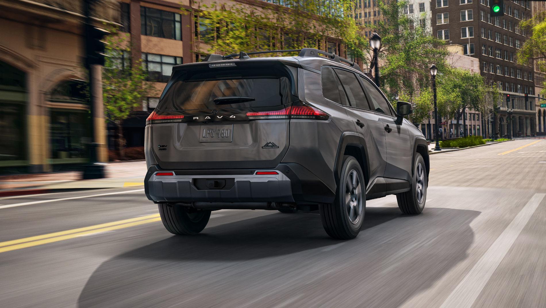 A gray Toyota RAV4 SUV drives down an empty urban street lined with trees and tall buildings on a sunny day.