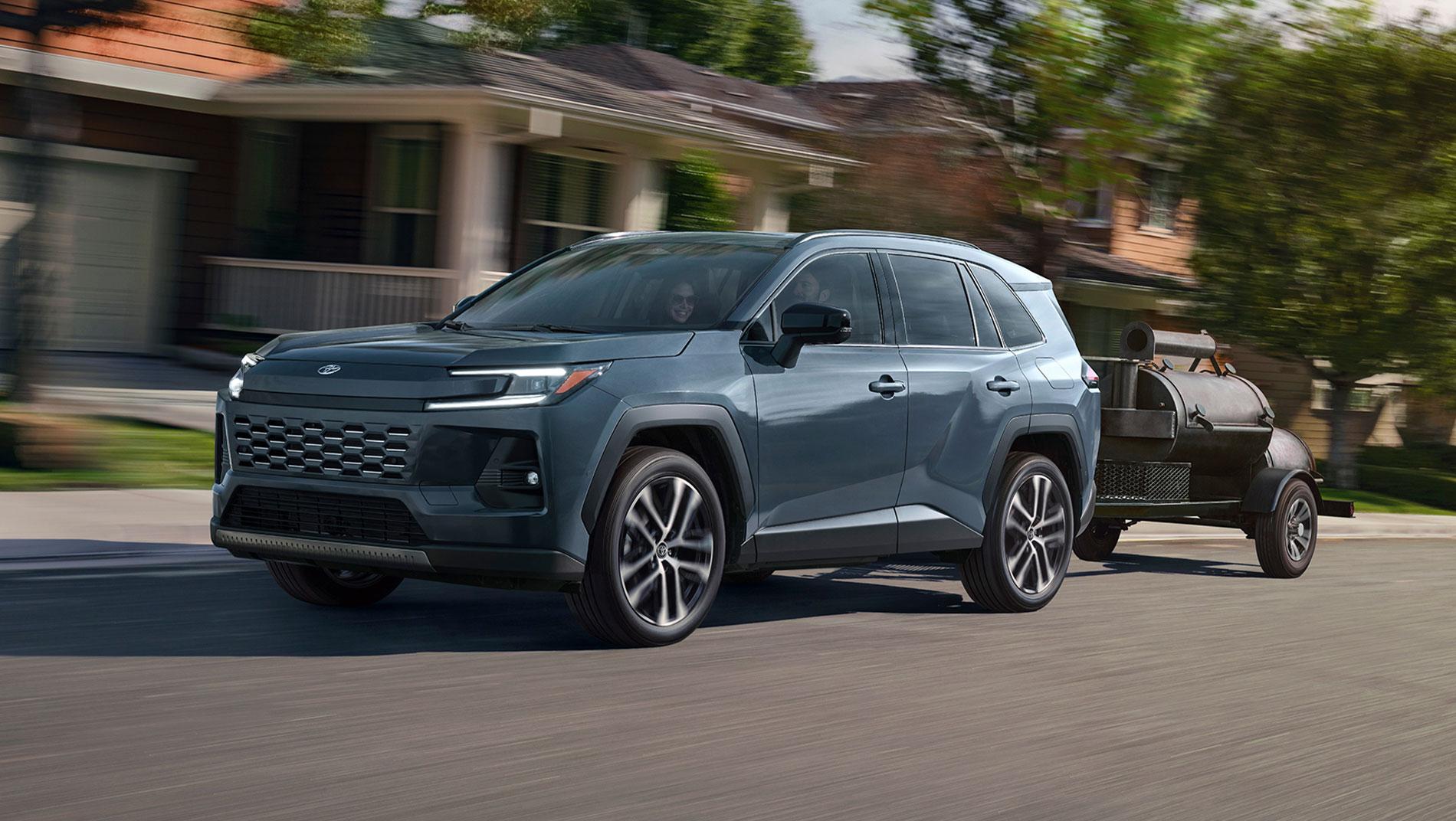 A gray SUV drives down a suburban street, towing a small black trailer with equipment. Houses, greenery, and a blurred background indicate the vehicle is in motion.