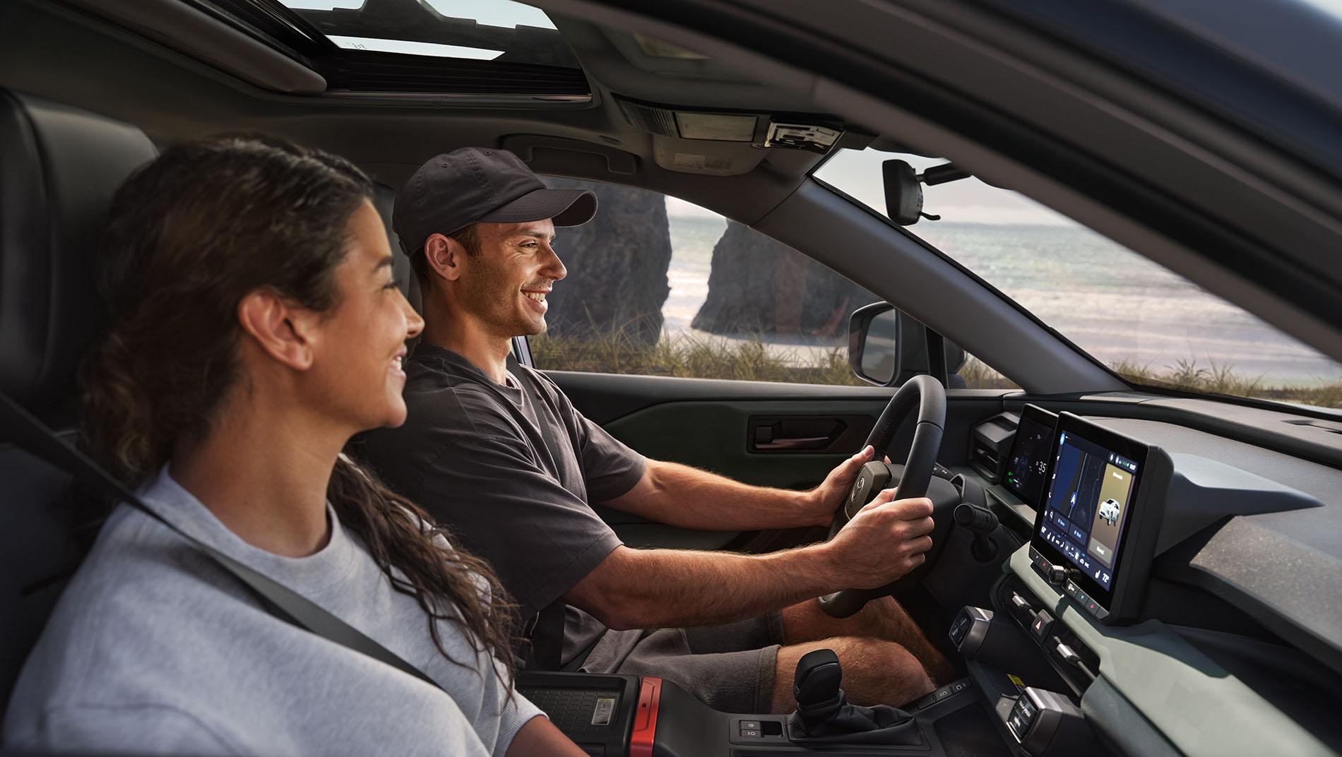 A smiling man and woman sit in the front seats of a car, driving along a coastal road with cliffs and the ocean visible through the windows. The man is driving, and both appear relaxed and happy.