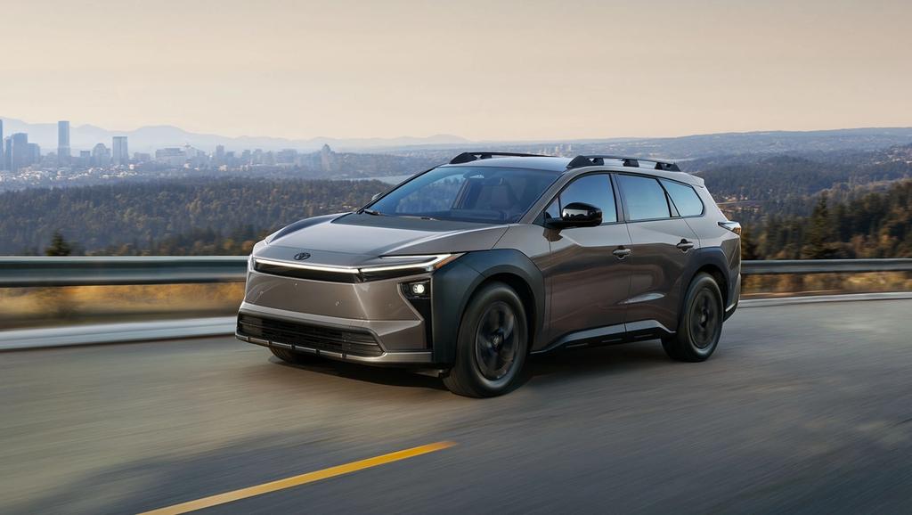 A modern gray SUV drives on a road with blurred motion, against a scenic backdrop of trees and a distant city skyline under a clear sky.