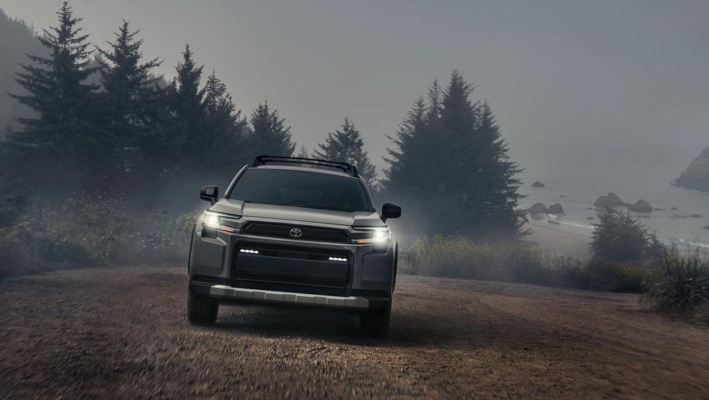 A gray SUV with headlights on is parked on a dirt road near misty pine trees and a foggy coastline, with the ocean partially visible in the background.
