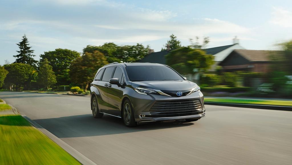 A gray 2025 toyota sienna is driving down a suburban street lined with trees and houses.