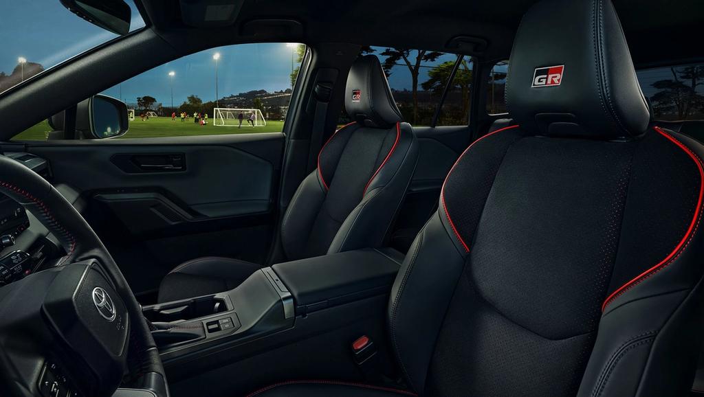 The image shows the interior of a car with black seats featuring red stitching and a GR logo. Through the window, a lit soccer field with players practicing in the evening can be seen in the background.