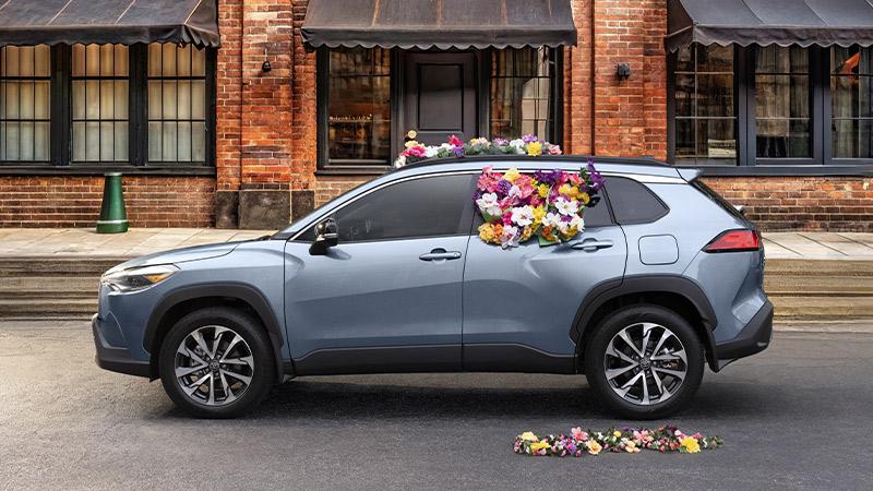 A blue SUV parked on a city street with colorful flowers overflowing from its open sunroof and scattered on the ground beside it. Brick buildings with awnings are in the background.