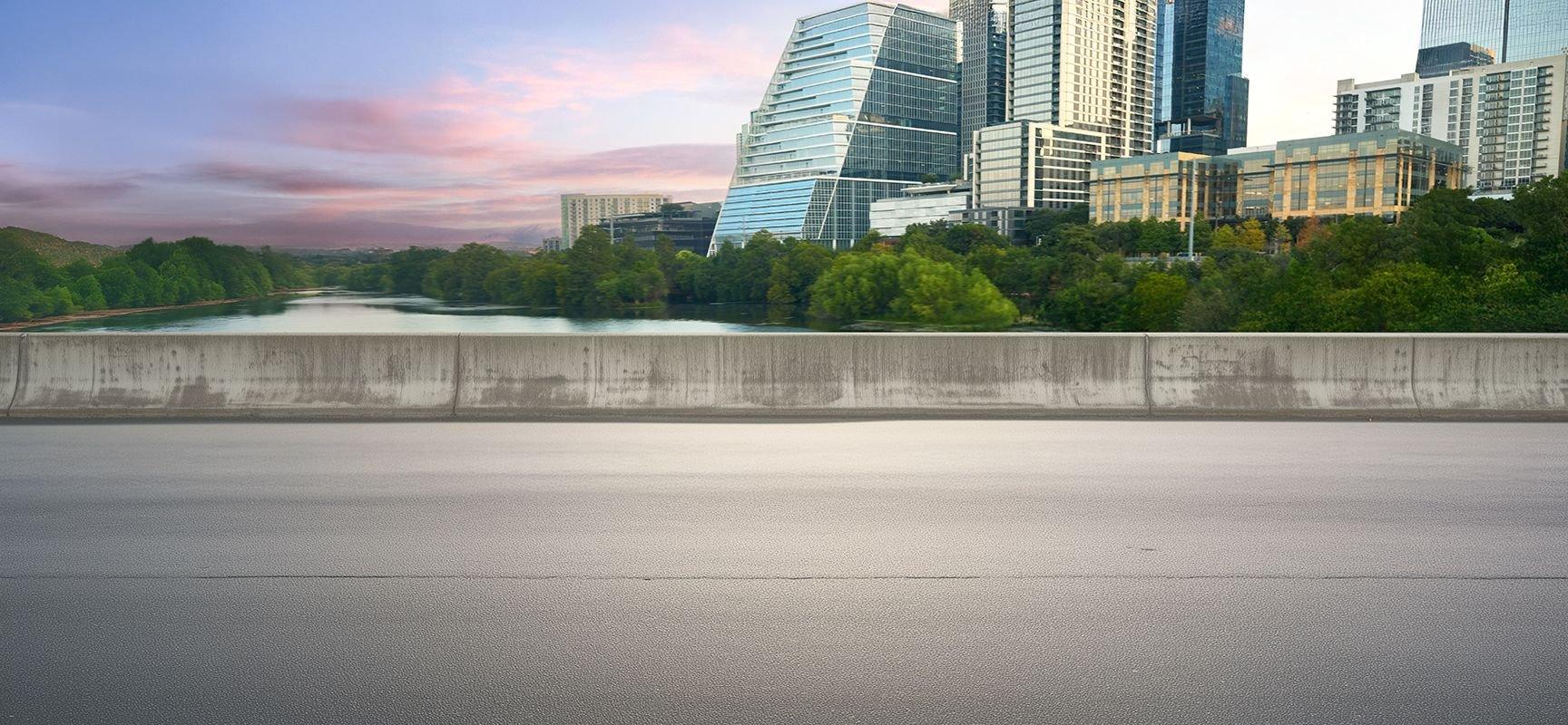 A cityscape with modern glass buildings and greenery reflected in a river, viewed from behind a concrete barrier on an empty roadway under a colorful sky at sunset.