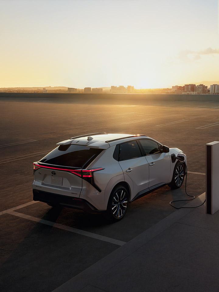 A white electric SUV is parked at a charging station on a rooftop parking lot at sunset, with a cityscape visible in the background.