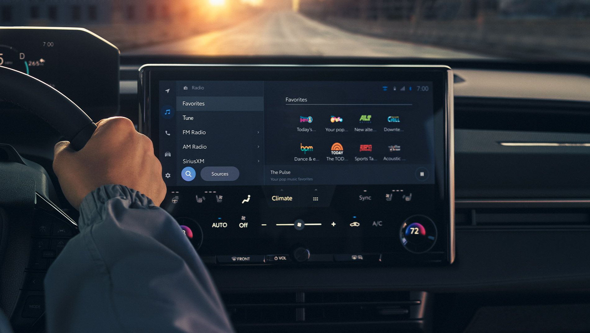 A person’s hand on a car steering wheel, with a close-up view of a large touchscreen infotainment system displaying radio and climate controls.