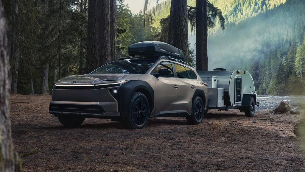 A modern SUV with a roof cargo box is parked in a forest, towing a small teardrop camper trailer. Tall trees and a river are visible in the background with mountains in the distance.