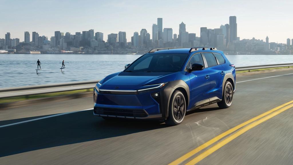 A blue electric SUV drives on a road beside a body of water, with a city skyline and two people paddleboarding in the background.