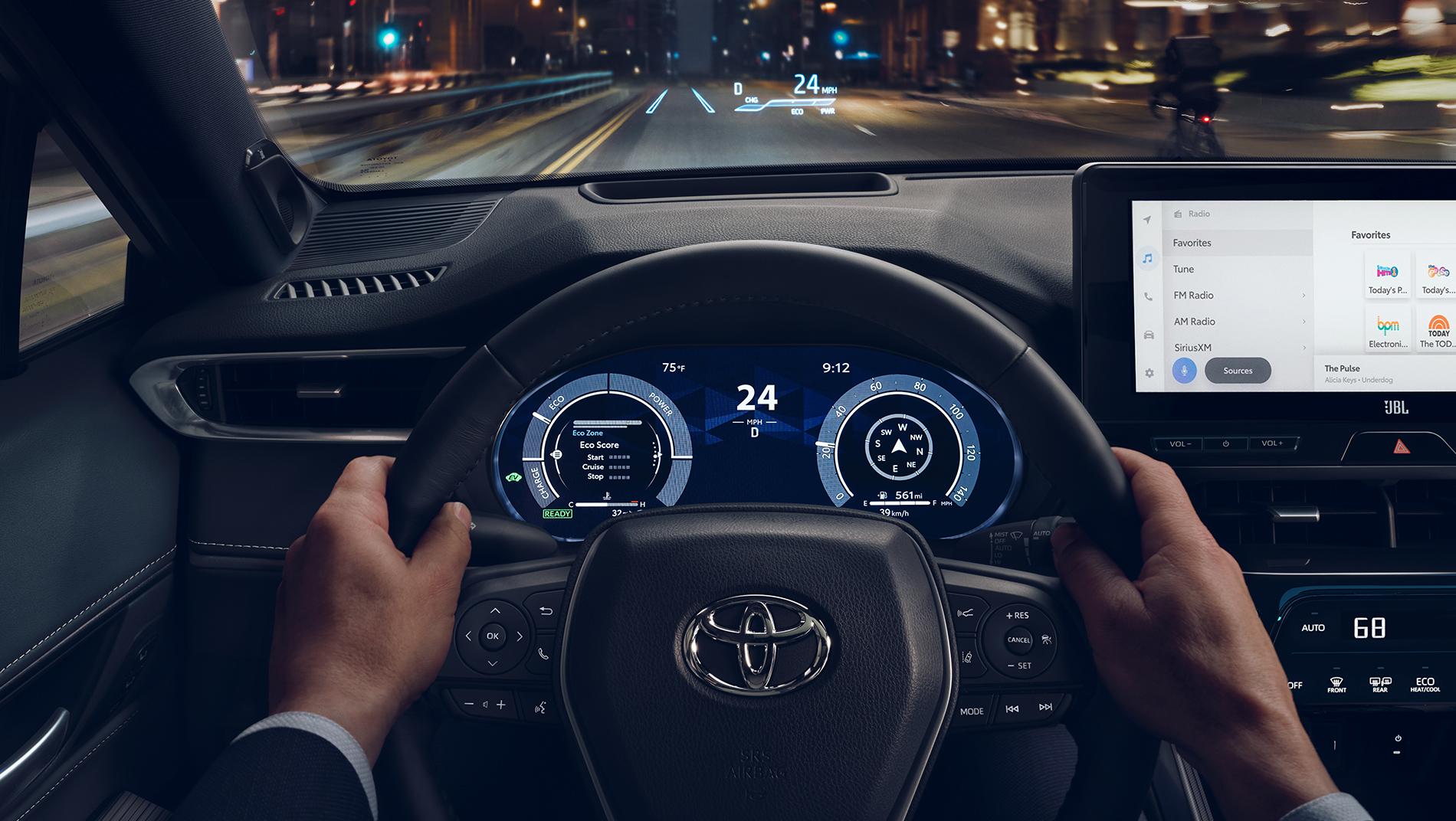 2024 Toyota Venza Hybrid inside looking out the front windshield driving down a city street at nightshowing touchscreen control panel, digital gague cluster, and heads up display showing important dashboard features on the windshield.