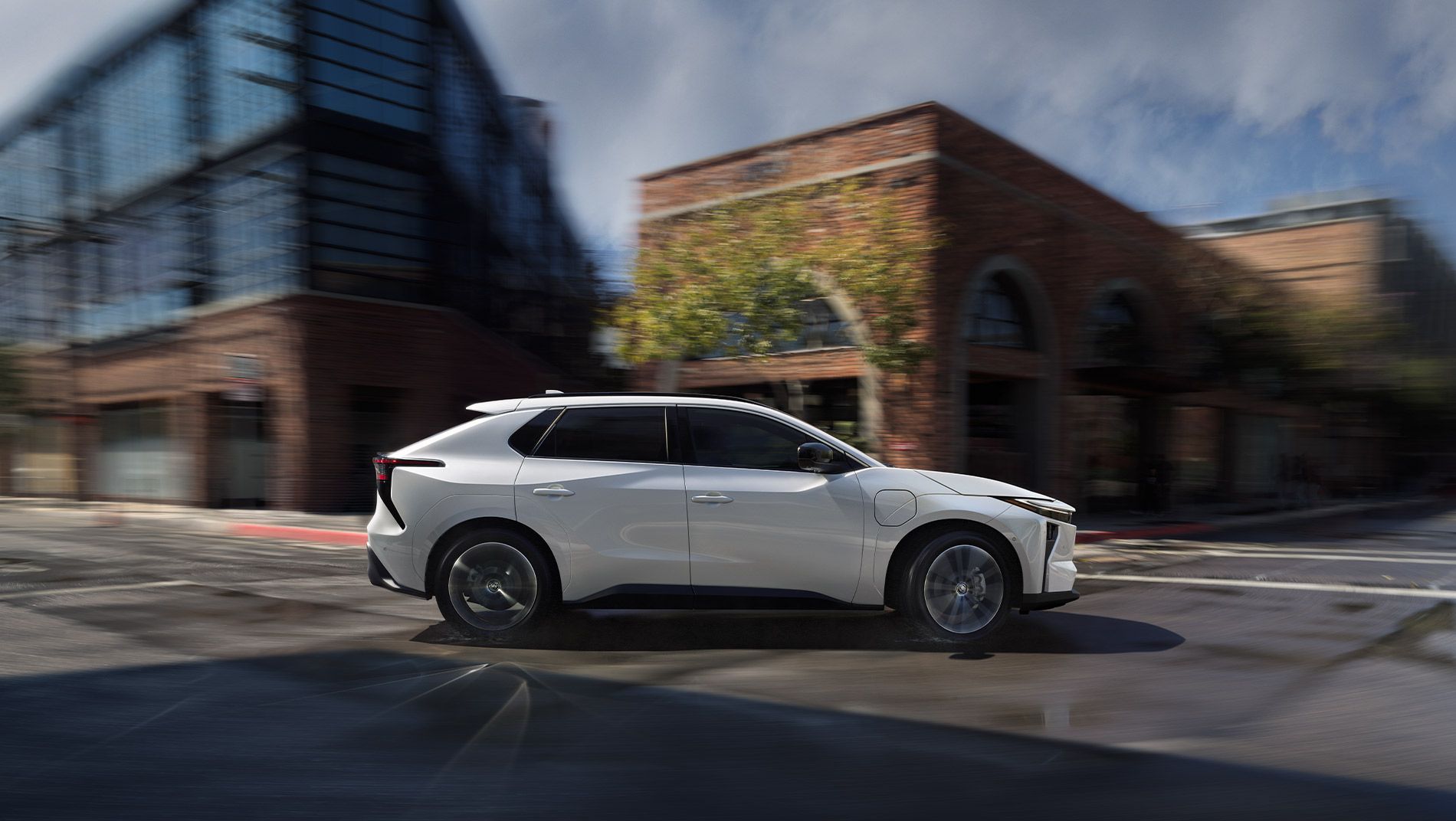 A white, modern SUV drives through an urban intersection lined with brick and glass buildings on a cloudy day.