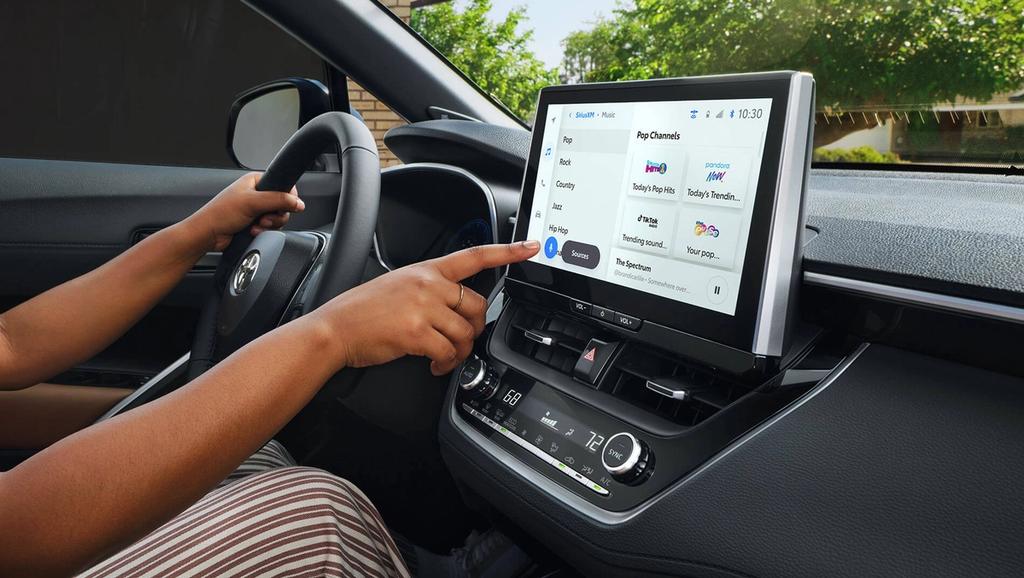 A person inside a car uses the touchscreen infotainment system on the dashboard, selecting music streaming options while holding the steering wheel with their left hand.