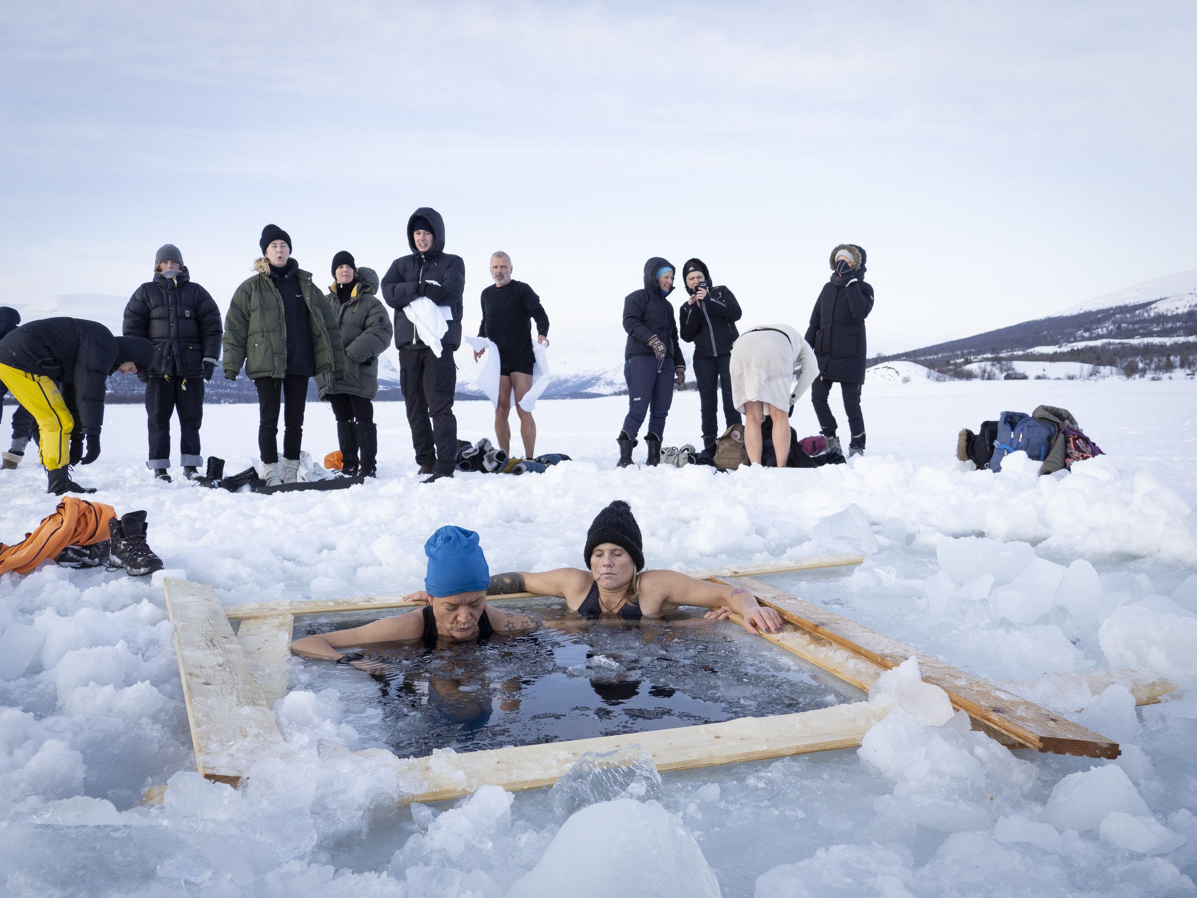 Two bathers in a square hole cut in the frozen lake, with a group watching from the ice