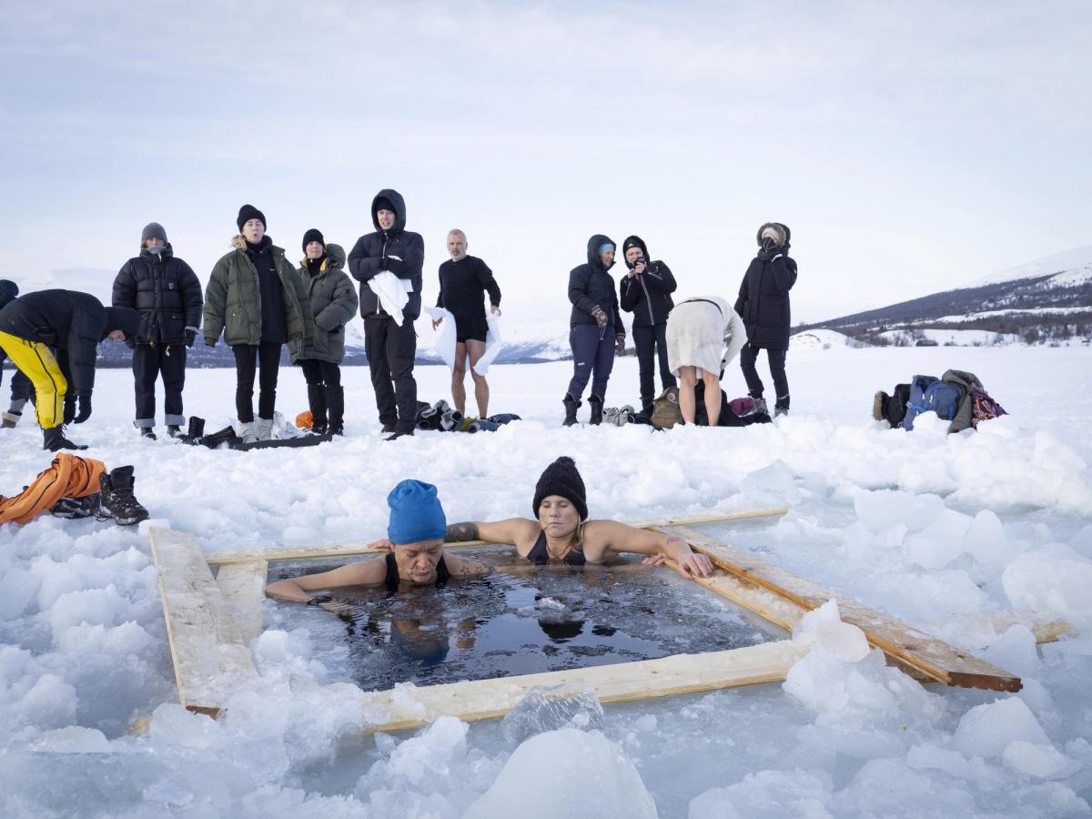 Two bathers in a square hole cut in the frozen lake, with a group watching from the ice