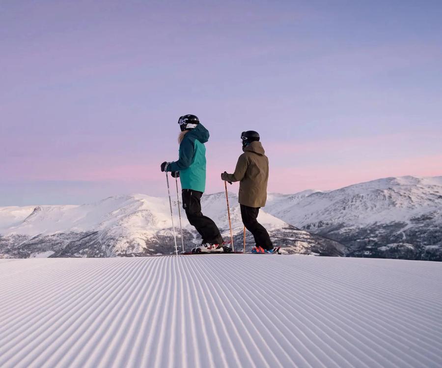 To skiløpere på toppen av en nypreparert bakke, med snødekte topper og rosa morgenhimmel bak seg