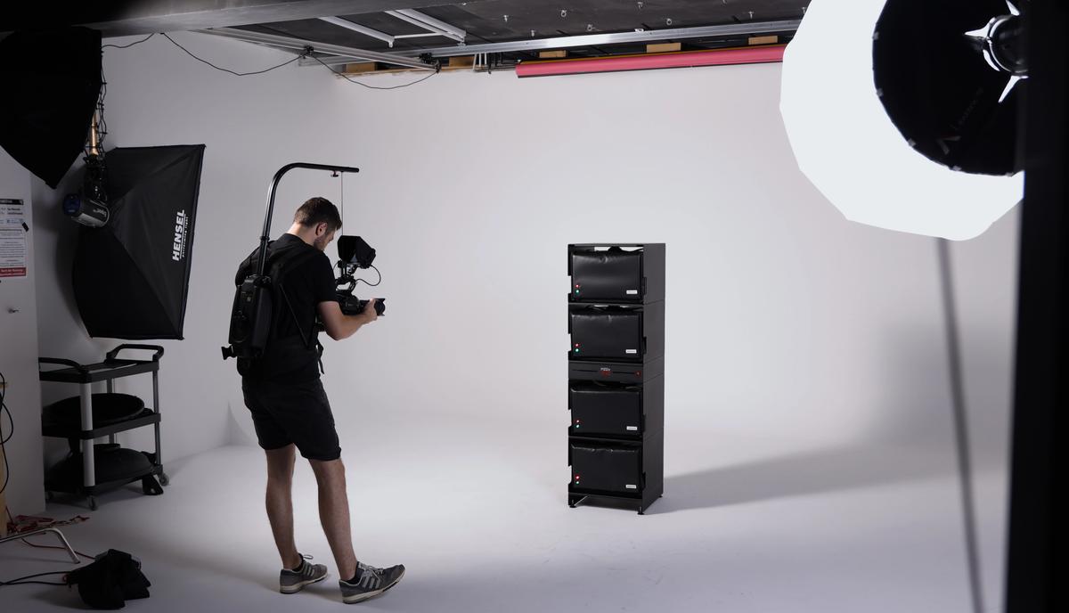 Ein Kameramann filmt im Fotostudio einen Stapel schwarzer Boxen, links und oben große Softboxen.