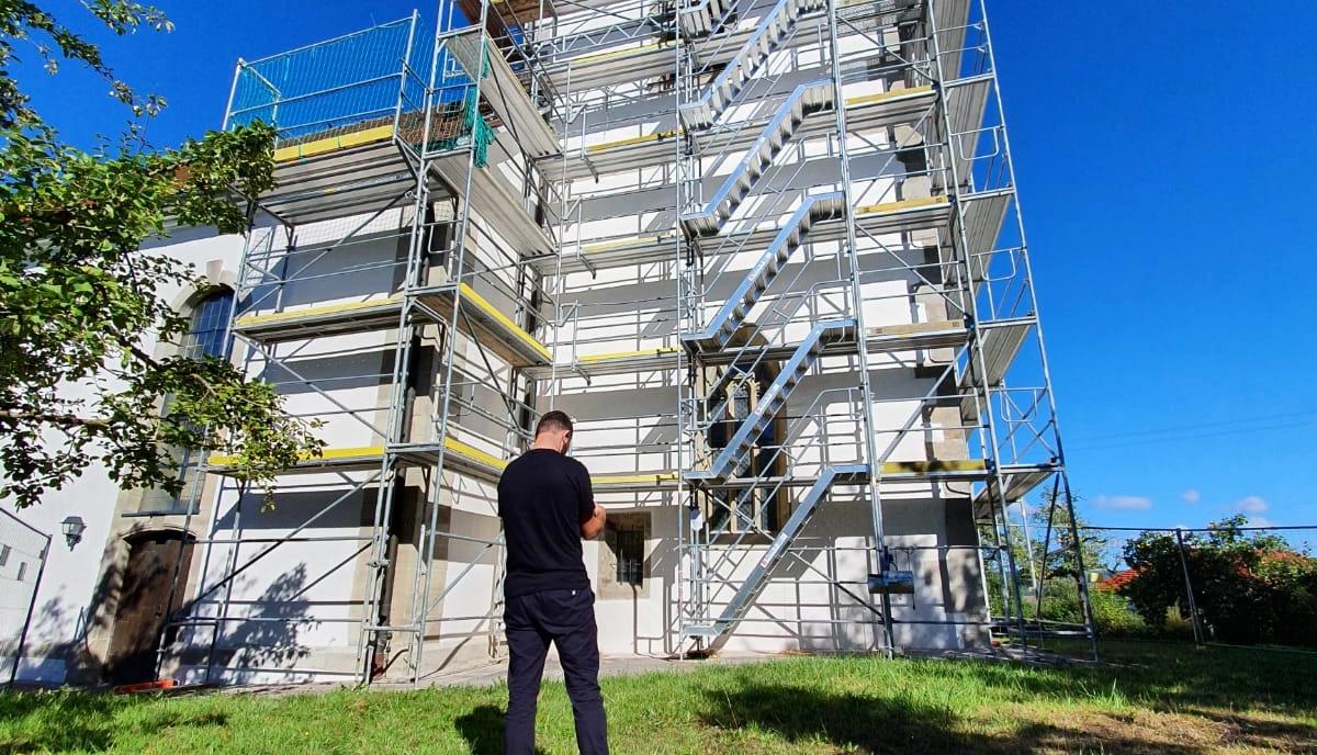 Ein vollständig eingerüstetes Gebäude unter blauem Himmel, davor steht eine Person auf der Wiese.