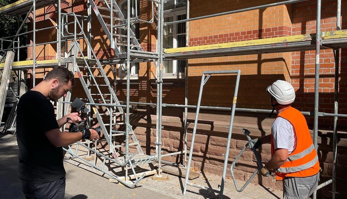 Ein Kameramann filmt einen Gerüstbauer mit Warnweste und Helm vor einer eingerüsteten Fassade.
