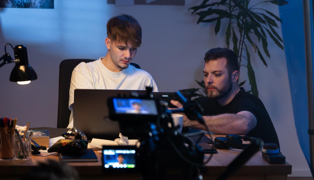 Zwei Männer arbeiten am Schreibtisch vor Laptop und Monitor, im Vordergrund die auf sie gerichtete Filmkamera.