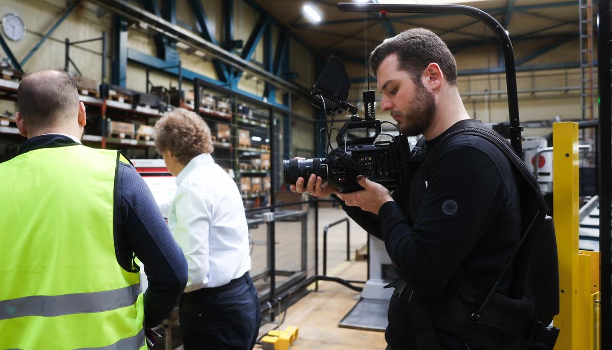 Werkstatt oder Lagerhalle, eine Person in Warnweste mit dem Rücken zur Kamera, eine zweite in dunkler Kleidung gegenüber, Regale im Hintergrund