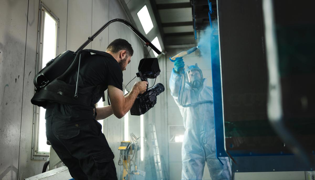 Kameramann filmt einen Arbeiter im Schutzanzug mit Spritzpistole in einer industriellen Lackierkabine.