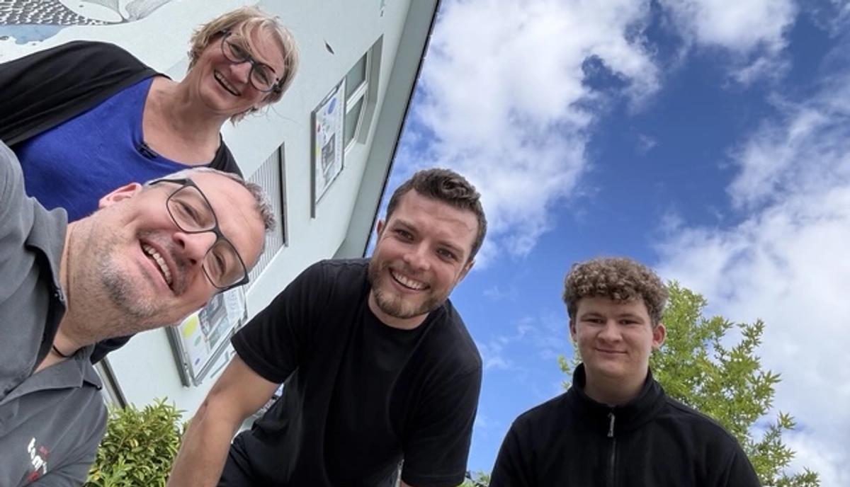 Gruppenselfie: vier Personen stehen dicht beieinander und lächeln in die Kamera, dahinter blauer Himmel.