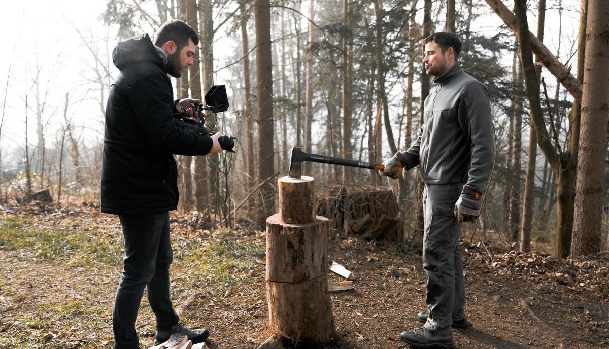 Ein Kameramann filmt im Wald einen Mann neben einem Hackklotz mit gespaltenem Holz.