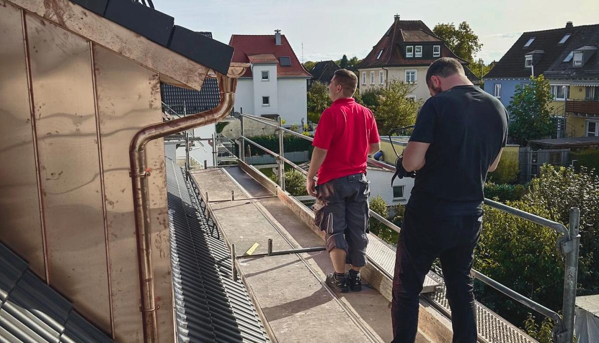 Ein Kameramann filmt auf dem Ziegeldach einen Handwerker im roten Shirt, dahinter Wohnhäuser und Bäume.