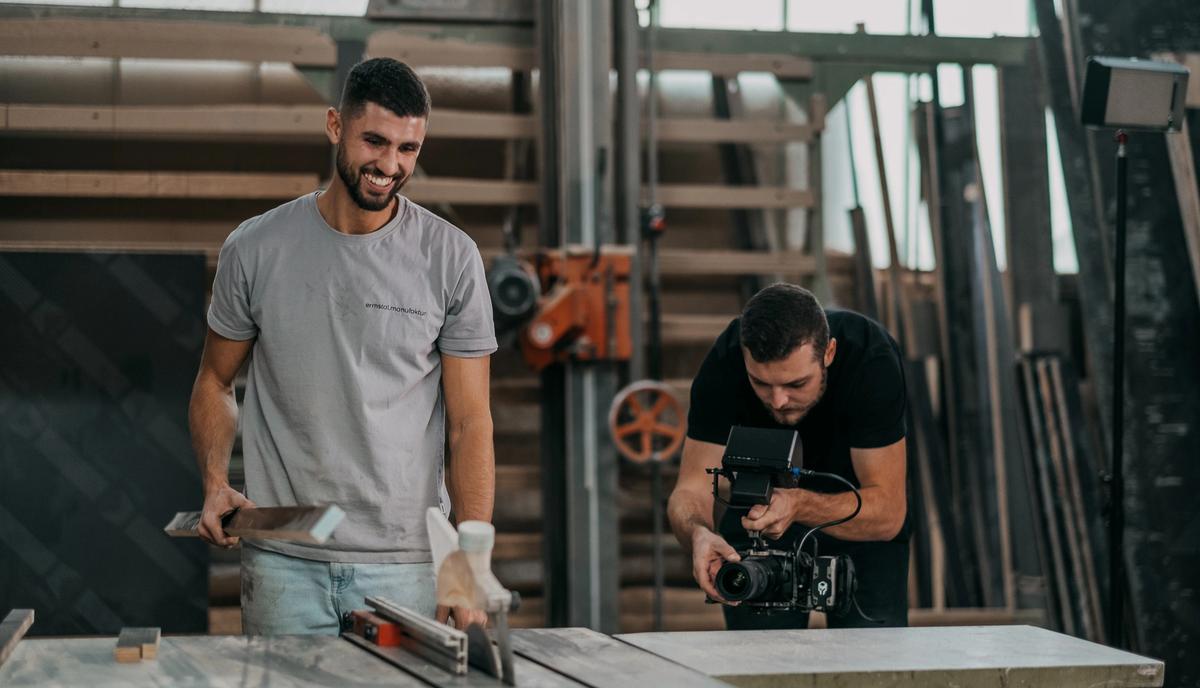 Ein Handwerker mit Arbeitshandschuhen arbeitet an der Werkbank, dahinter filmt ihn ein Kameramann.