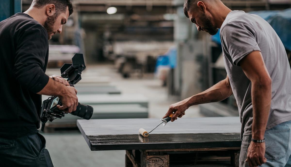 Ein Kameramann filmt von links einen Handwerker, der an einem großen Metalltisch ein Werkstück bearbeitet.
