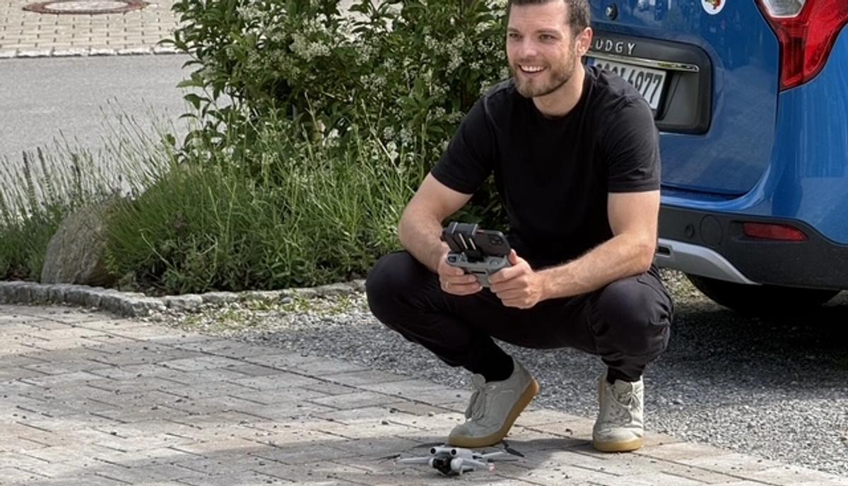 Ein Kameramann kauert mit seiner Kamera auf einer gepflasterten Einfahrt, dahinter ein blaues Auto.