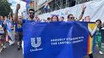 A group of people from Unilever walking and waving flags at Pride London, whilst the people at the front hold a Unilever sign with text saying 'Proudly Standing with the LGBTQI+ Community'