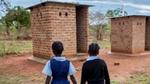 Two school children approaching a red brick toilet building.