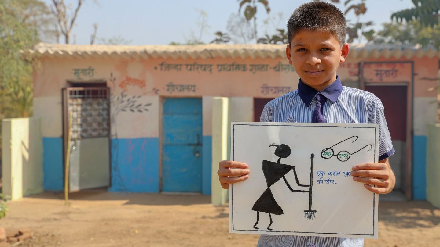 A schoolboy stands in front of a block of toilets holding a hand-drawn poster