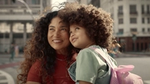 A woman holds a girl in her arms and both gaze in the same direction with smiles on their faces. Both have curly brown hair.