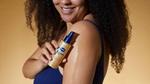 Woman with curly hair holding Vaseline Radiant X Replenishing Hydrating Body Oil spray. The product is on her arm. 